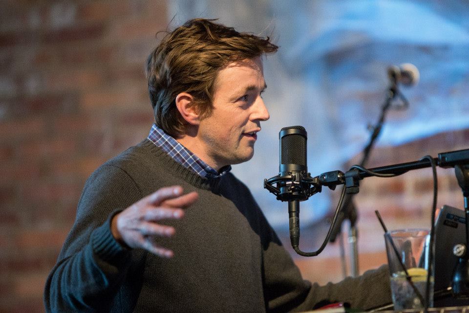 The background is neutral red brick. A white man with brown hair talks into the micrphone. He is wearing a grey sweater with a blue plaid collar. He has one hand raised as he talks.