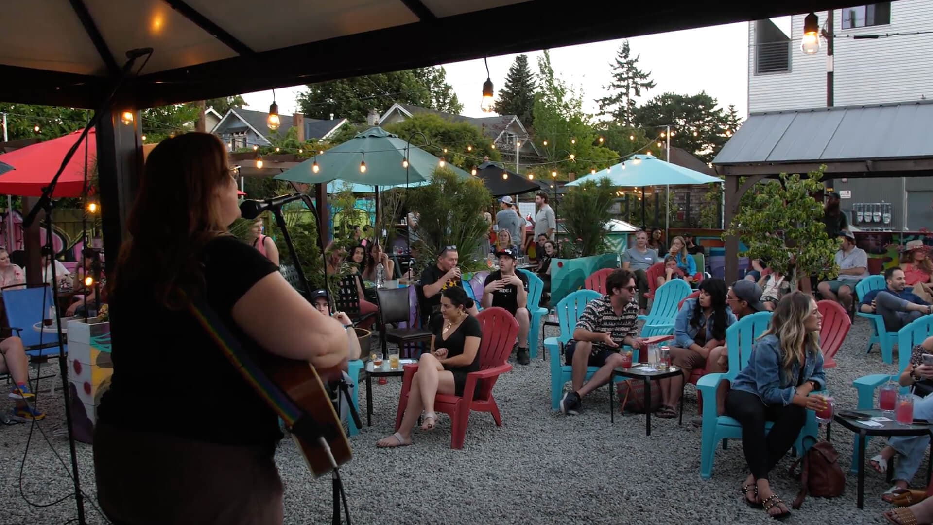 Image shows a woman playing guitar to a crowd outside at the Shady Pines Radio open mic