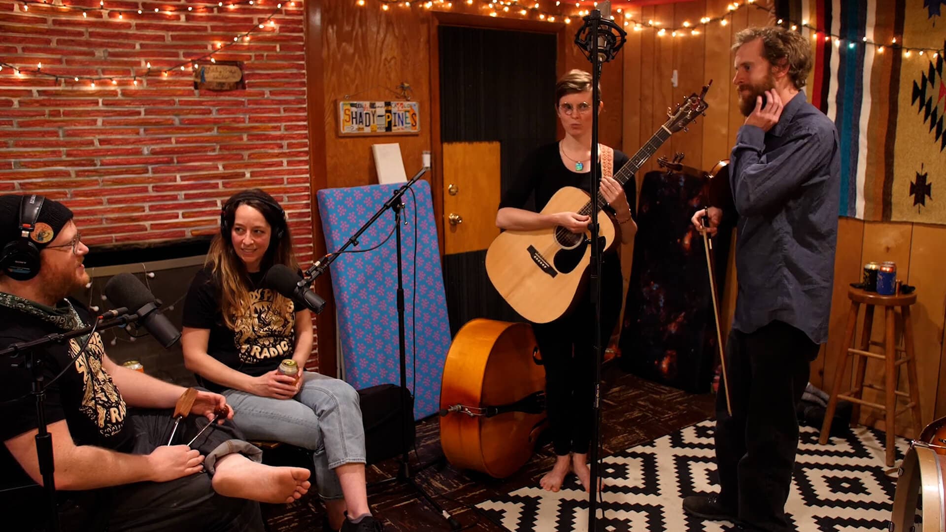 Image shows the Shady Pines Radio band in session. Two instrumentalists are standing up and two singers are sat on a sofa.