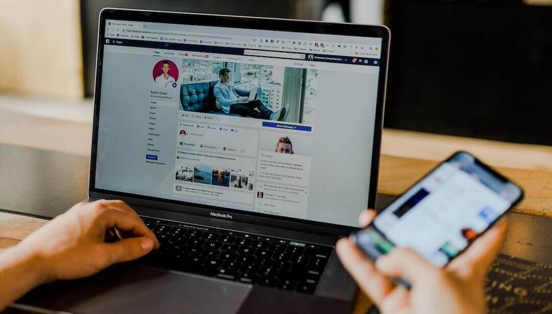 This is a photo of someone using social media on a Macbook in one hand, and their other hand is holding up a phone on social media. The background is neutral.