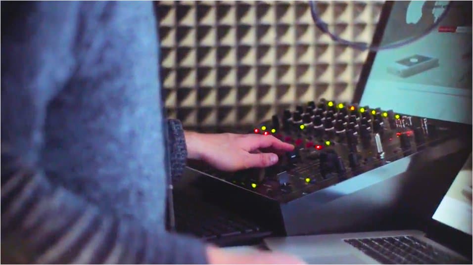 Image shows a person in standing in a radio booth operating a broadcasting desk and using a laptop.