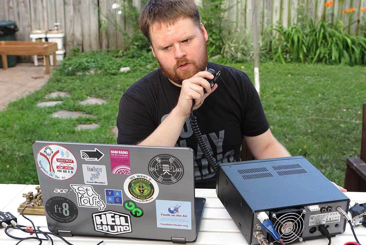 Image shows a man sitting in a garden, with a makeshift radio studio. He is speaking into a handheld microphone, and looking at a laptop.