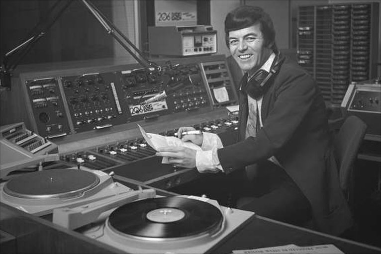 This is a black and white image of Tony Blackburn in the 70's DJing in BBC Studios. He is a white man with black hair and a big smile. He is wearing a suit and holding a piece of paper and a pen. He sits in front of a mixing desk and a vinyl player.