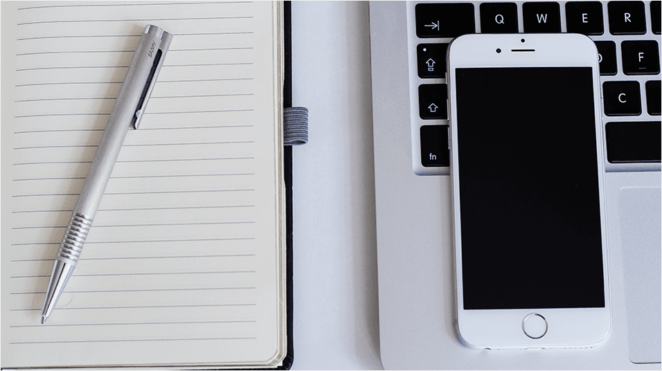 This is an image of a white lined notepad with a silver pen on top. On the right is a Macbook with just the keys visible, and a white iPhone on top.