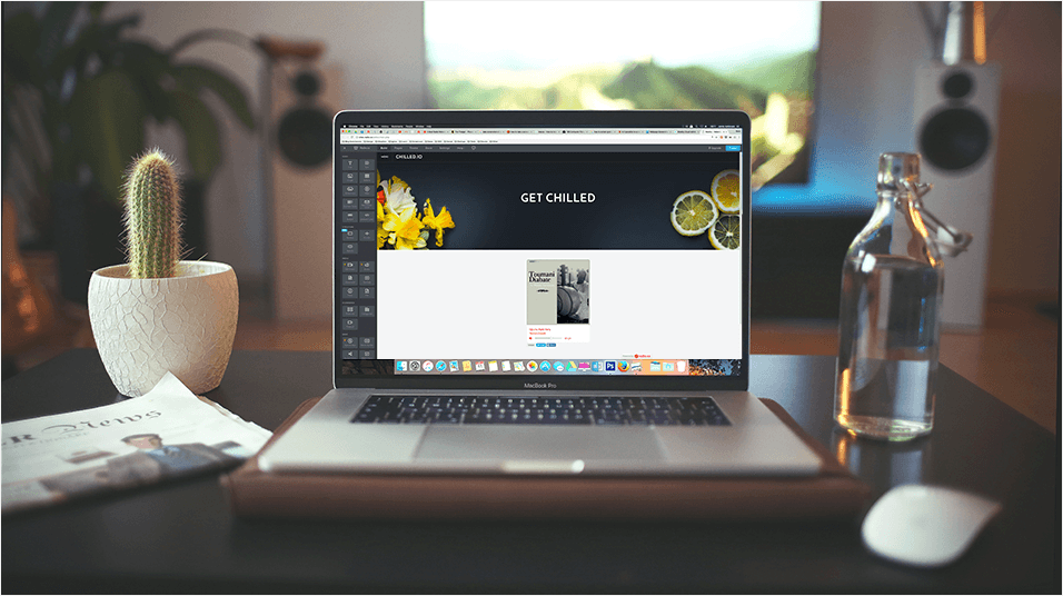 This is a picture of a Macbook on a table. On the left is a cactus in a white pot. On the right is an open glass bottle of water. In the background is a mic on a stand.
