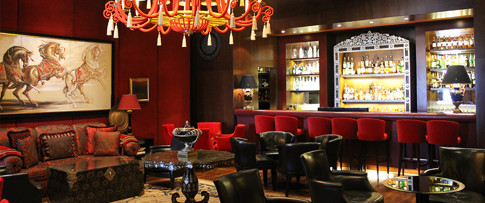 This is an elegant red hotel bar, with a red chandelier in the centre of the image. On the left there is a framed picture of horses. Along the bar there are a set of red seats and a drinks cabinet.