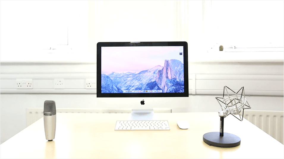 This is an image with a white backdrop with a silver Mac in the centre. The Mac's screen is a generic purple background. On the left is a silver Mic, on the black is an angular black lamp.