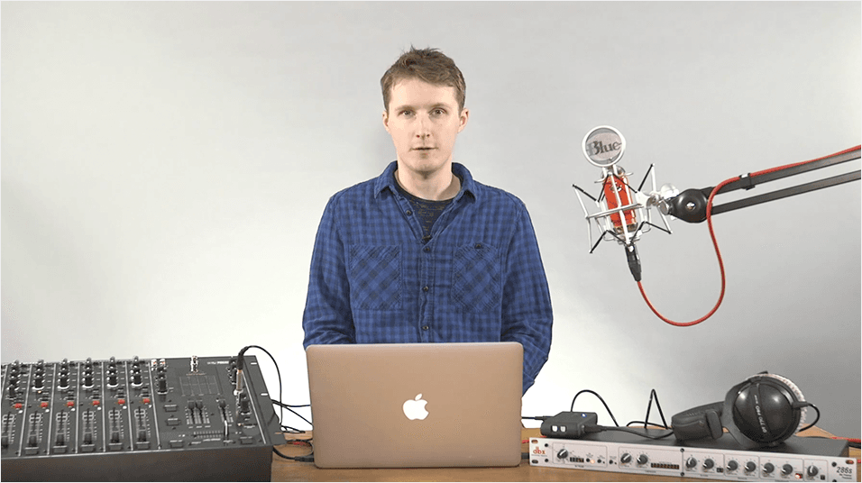 This is an image of a white man with short sandy blonde hair, looking into the camera and wearing a blue shirt. He is stood in front of a Macbook and speaking into a microphone on an arm. On the left is a black mixing desk on the right is a keyboard.