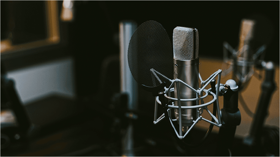 There is a black backdrop of a radio studio. There is a black mic on a stand visible in the centre of the image, with triangular elements visible.