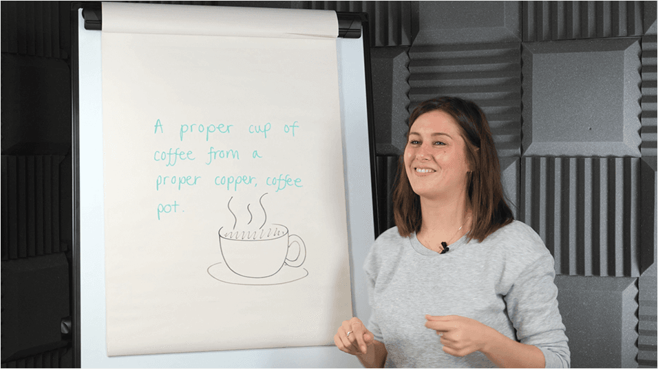 A white woman with shoulder length brown hair smiles in front of a whiteboard with advice on voice warm up exercises on it.