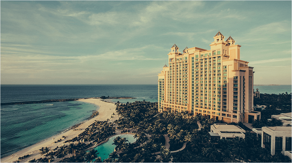 This is an image of a large beachside 5 star hotel. The sea is on the left of the image with the coast in azure blue, and the tower on the right is gold from the sunlight. The sky is as blue as the sea and meets it midway through the image.
