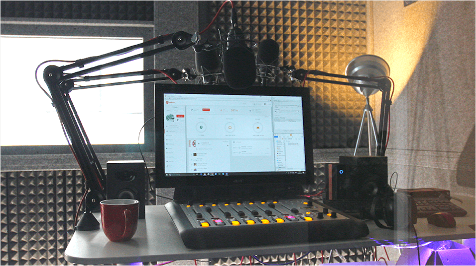 This is an image of a small home studio. In the background is warm light and soundpanels. In the foreground of the image is a monitor with a mixing desk in front of it.