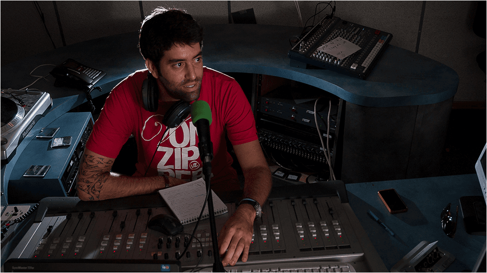 This is an image of a man with black hair and a black beard, sat in front of a mixing desk and a green microphone. He is presenting radio. The background is a blue curved desk typiacl of a studio setup.