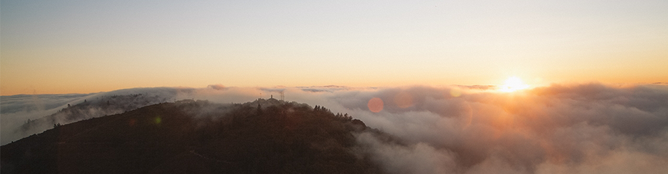This in image of the dawn of the sun over some clouds. The sky is a soft orange and the hills are dark and rolling.