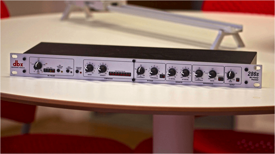 This is an image of the Dbx 286 S Microphone Processor, side on. It is a slim grey panel with various black knobs on it, on a white desk.
