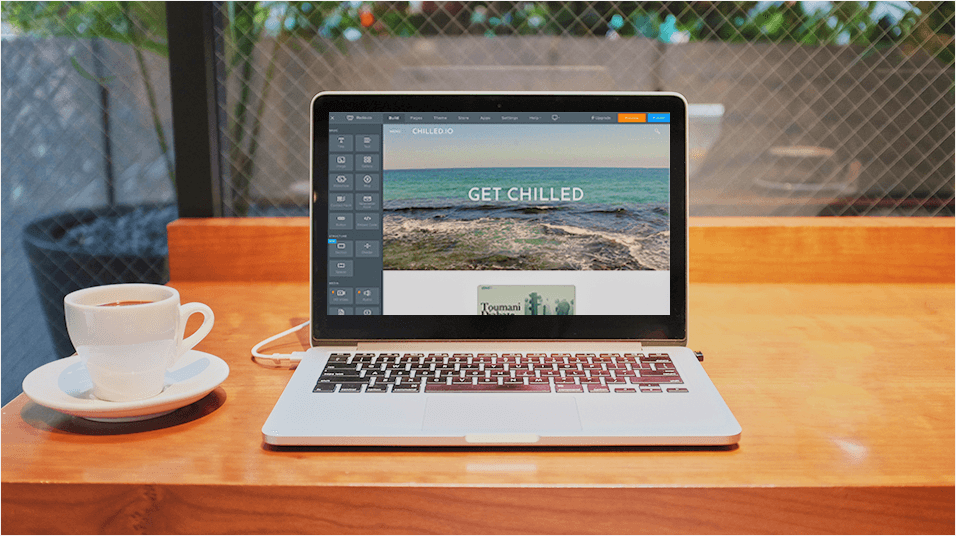 There is a silver and black Macbook in the middle of the screen. To the left of the Mac is a white coffee cup and saucer. The Mac is on a brown desk.