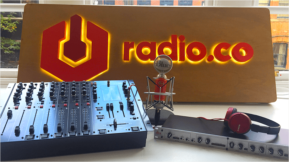 This is an image of a silver mixing desk next to a small silver and red mic and a pair of red and black headphones. There is a red and brown lit board with the radio.co logo visible.