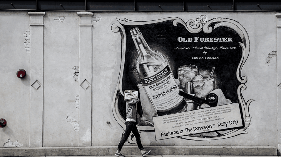 This is an image of a man dressed in black walking in front of a large white billboard/mural. it is advertising Old Forester with an image of a bottle.