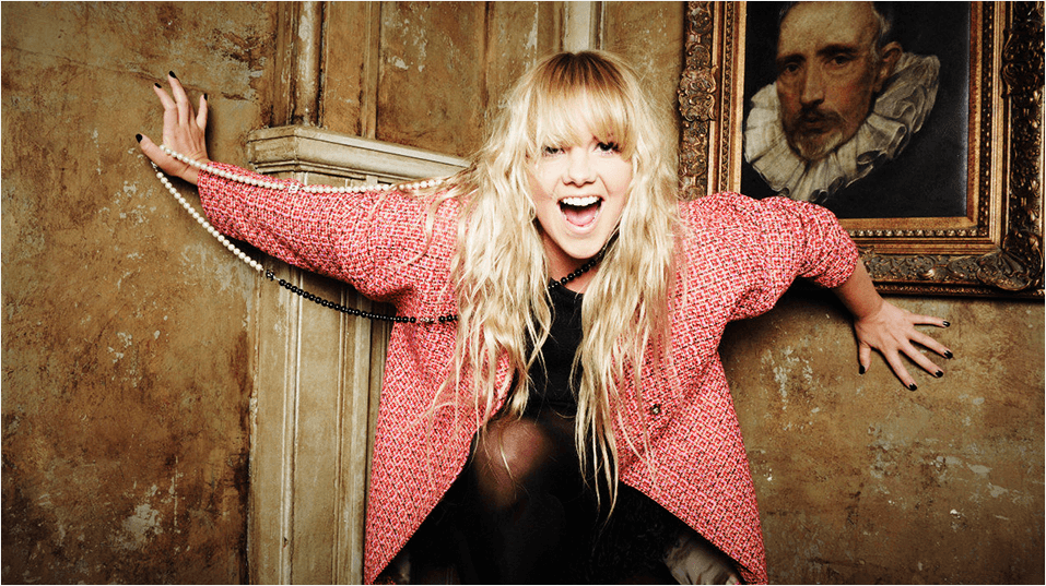 This is a picture of Goldie Rocks, a white woman with long blonde hair and a straight across fringe. She is laughing with her mouth open, with her arms against a wall. Above her is a framed picture of (maybe?) Francis Bacon in a rruff.