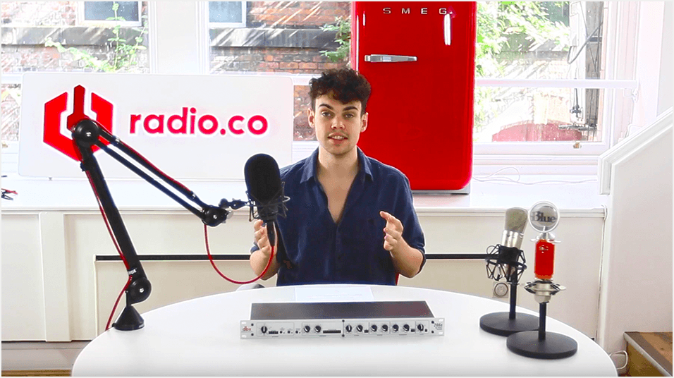 This is a picture of a white man with black hair and a blue shirt sitting in front of a microphone on a boom arm and small mixer. He is pointing at the mic. The back of the image is a red radio.co logo on a white sign.