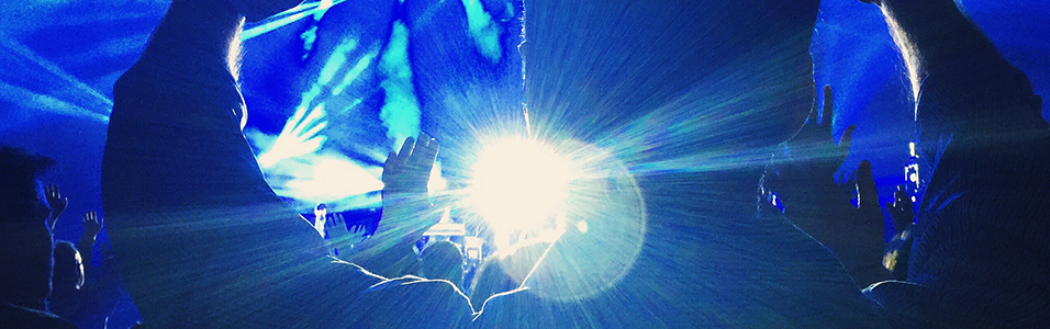 This is a dark blue image that appears to be from a club or a concert. The laser lights are green and we can see silhouettes of dark figures with their hands up. There is a white lens flare in the centre of the image.