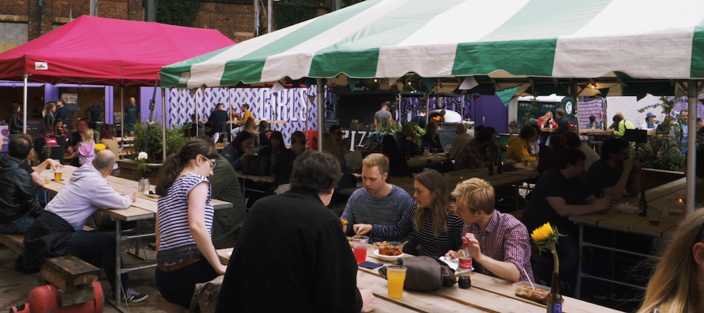 This is a screenshot of market stalls and food stalls at GRUB. There are two stalls in the image, a hot pink one and a green and white striped one. Beneath it are two long picnic tables and from behind are images of people eating and drinking.