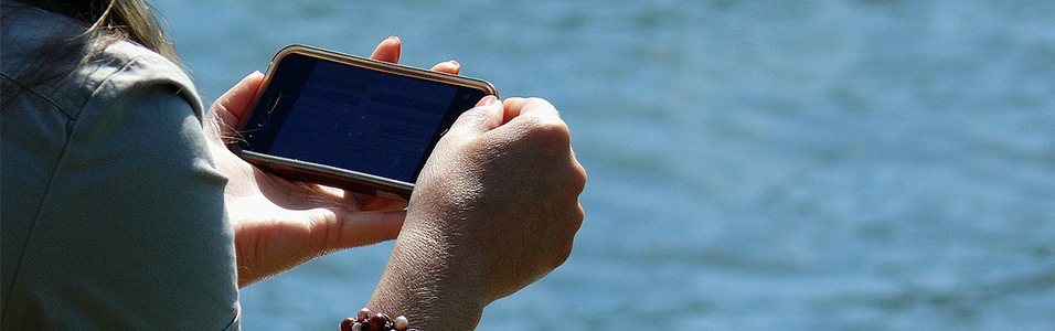 White hands against a sea background hold a blank black iPhone horizontally.