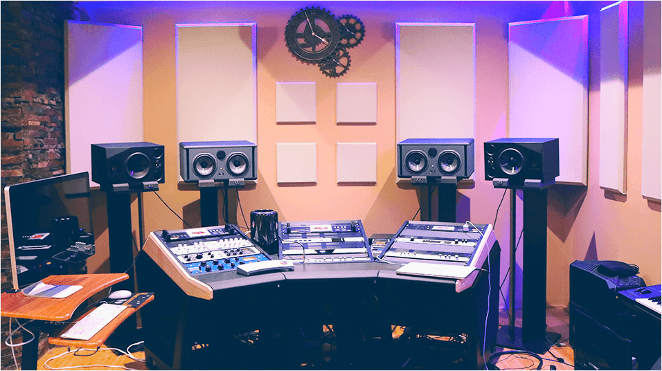 this is an image of a small radio studio, taken from behind. The forefront of the image is a big black mixing desk with speakers above and either side of it.