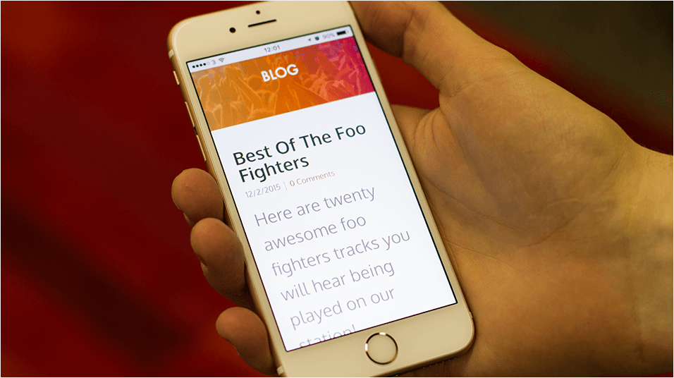 This is an image of a white iPhone in a hand against a red neutral backdrop. The phone has an orange banner and reads "BLOG' and below the blog topic is 'Best of the Foo Fighters' with a stock example of a blog article.