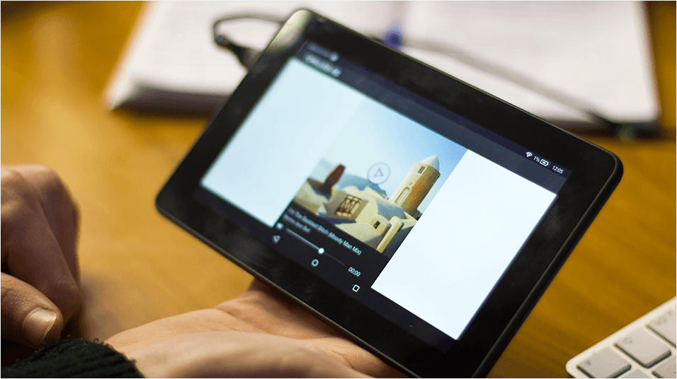 Against a wooden desk, there is a white open book visible. There is a black tablet open in the centre. On the screen is an indecipherable image of buildings with a black caption.