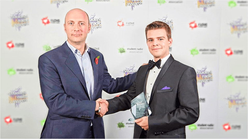 Two white men shake hands and the one on the left holds a glass award. The man on the right is bald and white, and the man on the left is younger and white with dark hair. they are both wearing suits.
