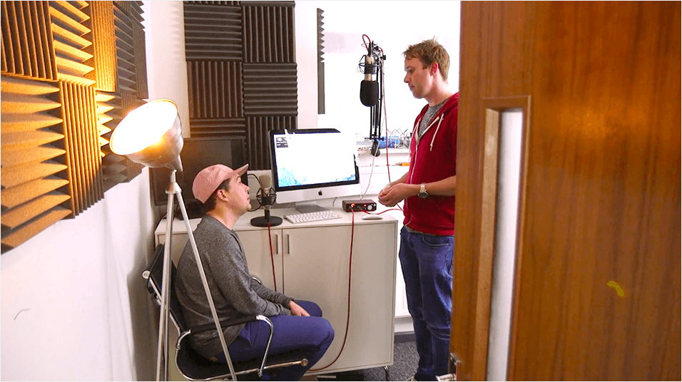 This is an image of a small radio studio. On the right is a brown tan door with a silver handle. One person sits in a grey shirt with a yellow cap. The other talks wearing a red jumper and blue jeans. There is a white lamp and yellow soundpanels visible.