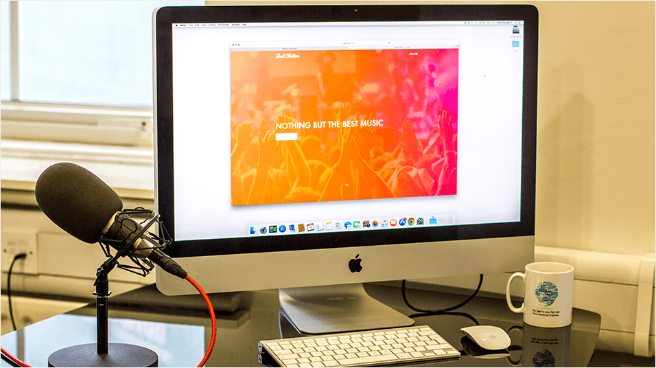 This is a black monitor with a white, pink and orange homepage visible with a taskbar at the bottom. It is a silver Mac. On th eleft there is a black microphone with red wiring.
