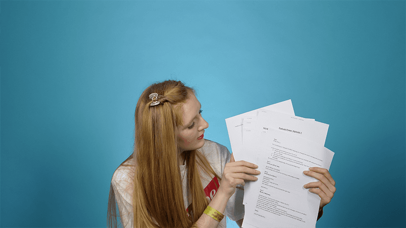 Against a plain blue backdrop, a white girl with long auburn hair holds three pieces of white paper in her hands - scripts.