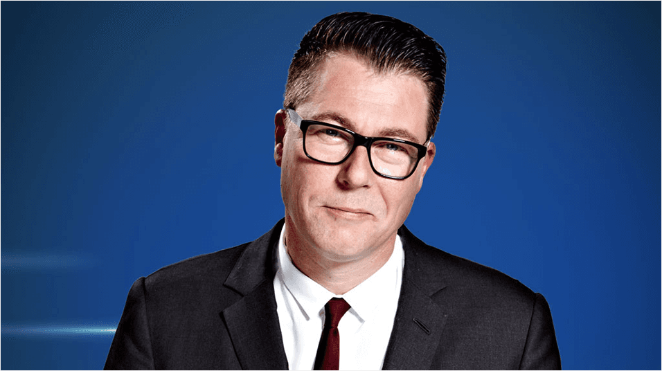 A white man with glasses and dark hair, Ian Collins, smiles at the camera. He is wearing a black suit and tie.