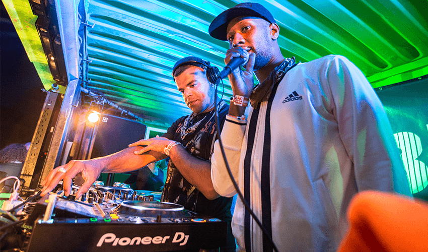 Image shows a male DJ and male MC performing at a music festival.