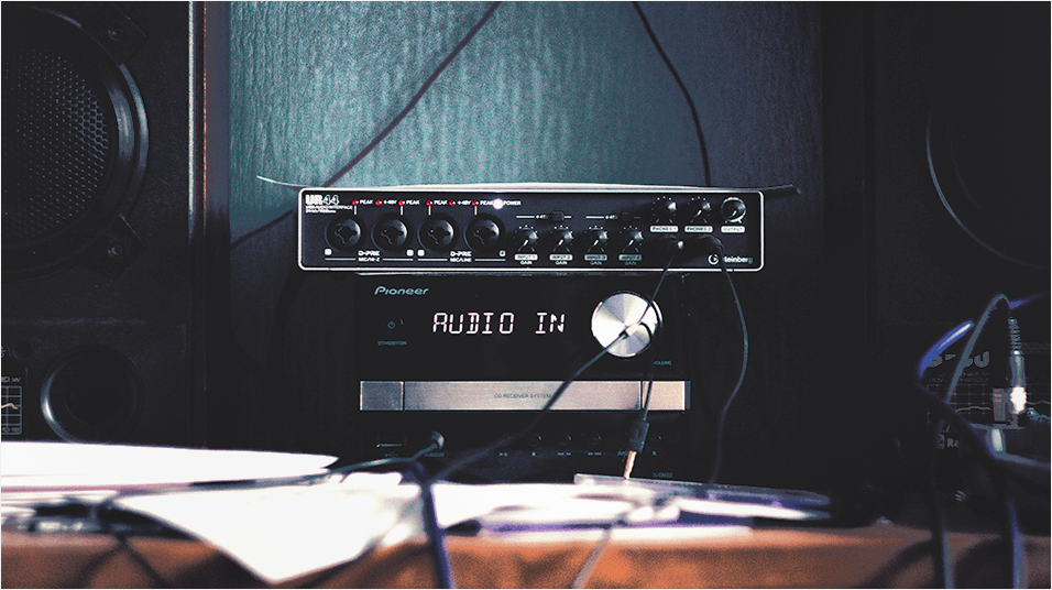 This is an image of a thin black audio interface. The details on it are silver. In the back there is a black speaker to the left of the image. The background of the image is dark blue.