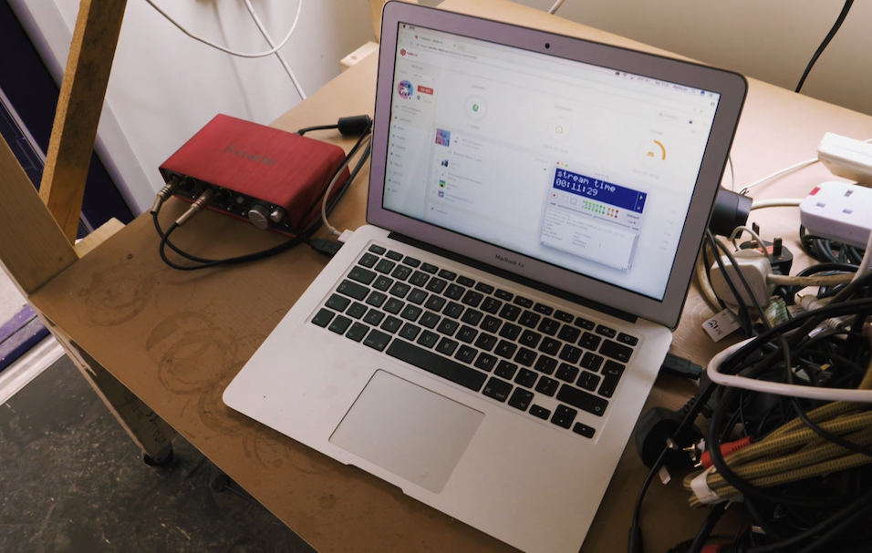 this is an image of a Macbook Air laptop in silver on a messy table. On the left, the interface is plugged into the Mac. On the right are tangled wires and extension leads.
