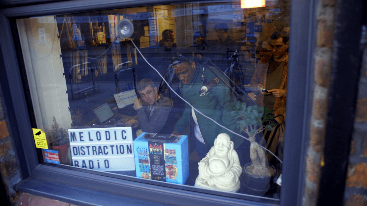 Image is a photo from the outside window of Melodic Distraction, two presenters can be seen broadcasting on the other side.