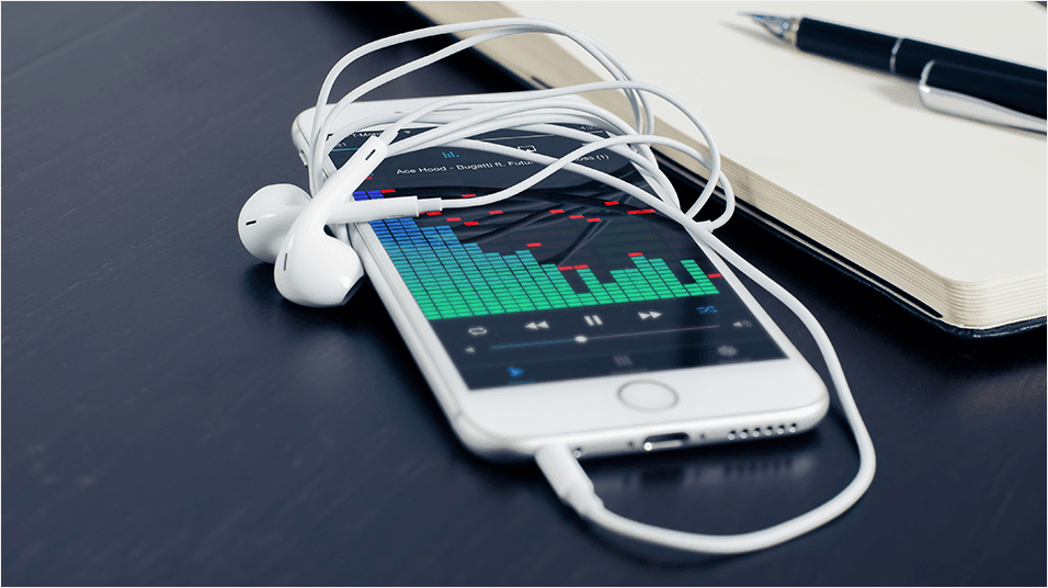 This is a black neutral backdrop with the corner of a Macbook visible. There is a white iphone in the middle with classic wired headphones connected and wrapped over the top. The screen reveals a green graph of sound levels in the middle.