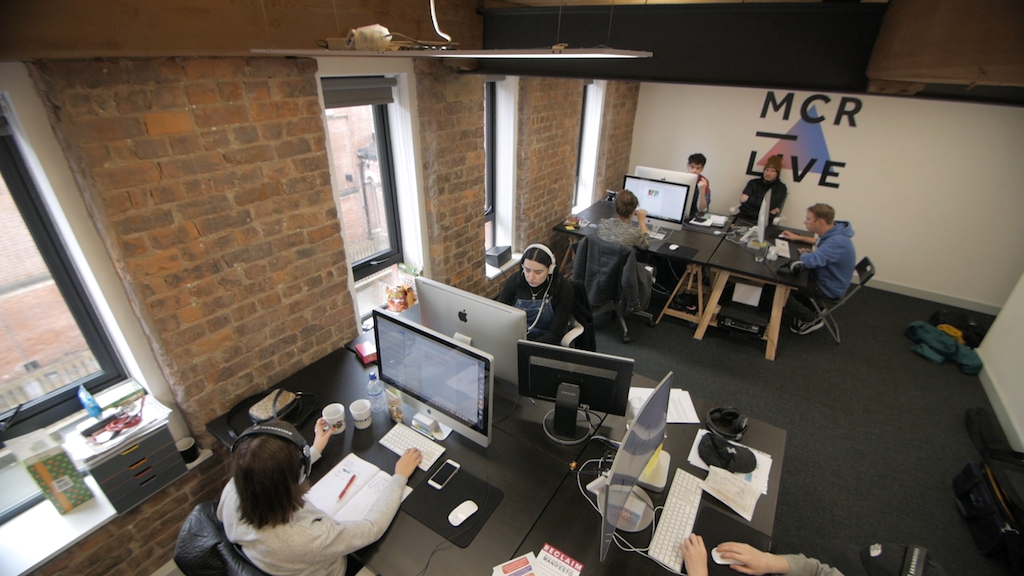 This is an image of the MCR Live Office. It is a bricked wall with two desks adjacent to each other. On these desks are 7 members of staff working at Macs. The back wall has the MCR Live logo on it (the words MCR Live over a blue and pink triangle).