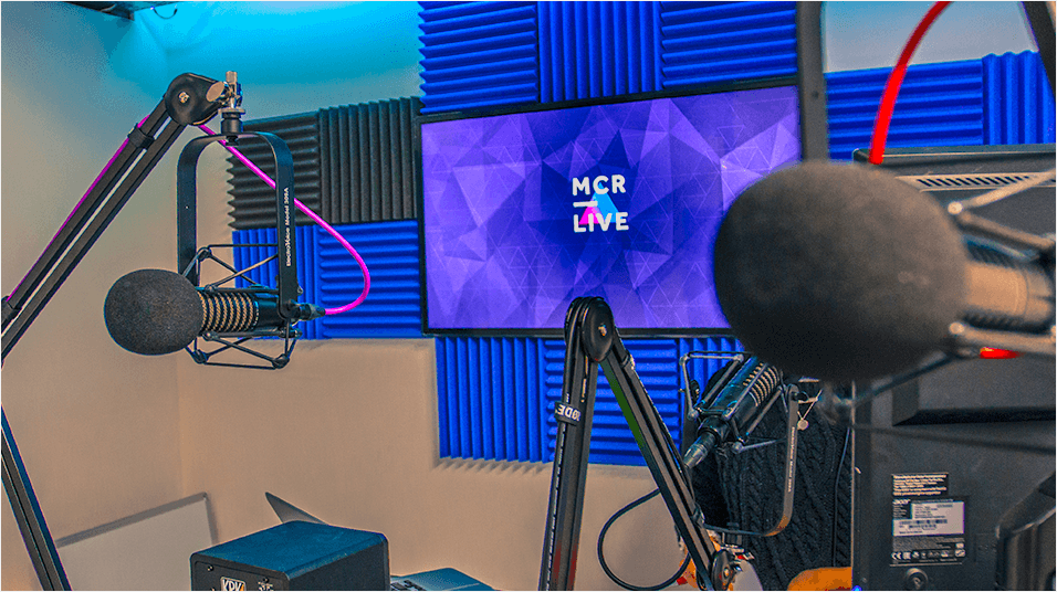 This is an image of the Mcr Live Studio. It has blue soundpanels in the background of the image and a blue monitor reading MCR Live. On the left is a mic on a boom arm, on the right is a mic on the desk. Both have red wires.