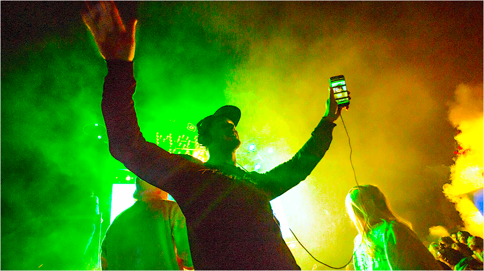 The background is green and yellow lighting in a haze. In the middle of the image is a man with a cap on raising his hands. His right hand has an iPhone plugged into a wire. He appears to be on stage.