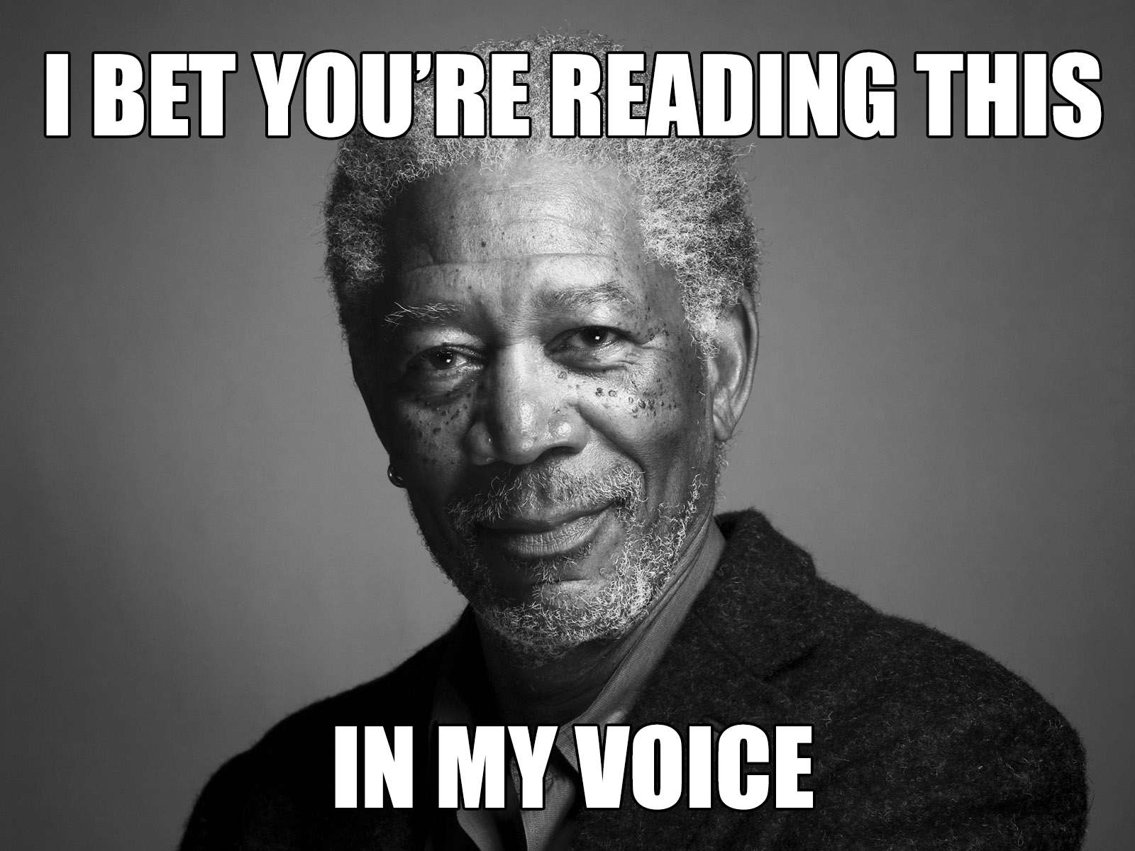 This is a black and white image of Morgan Freeman smiling at the camera. He is a black older man with grey hair, a grey beard and freckles. He is wearing a black shirt. Above him in classic 'Meme' font are the words I BET YOU'RE READING THIS IN MY VOICE