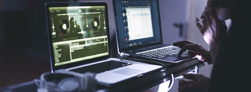This is an iamge of two monitors in the dark, presumably at night time. The first screen has a yellow mixer on. On the left is a list of tracks in white. There is a silhouette of someone with rings using the second laptop.