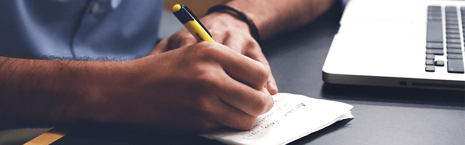 Image shows a close up of a man writing on a notepad, next to a laptop.