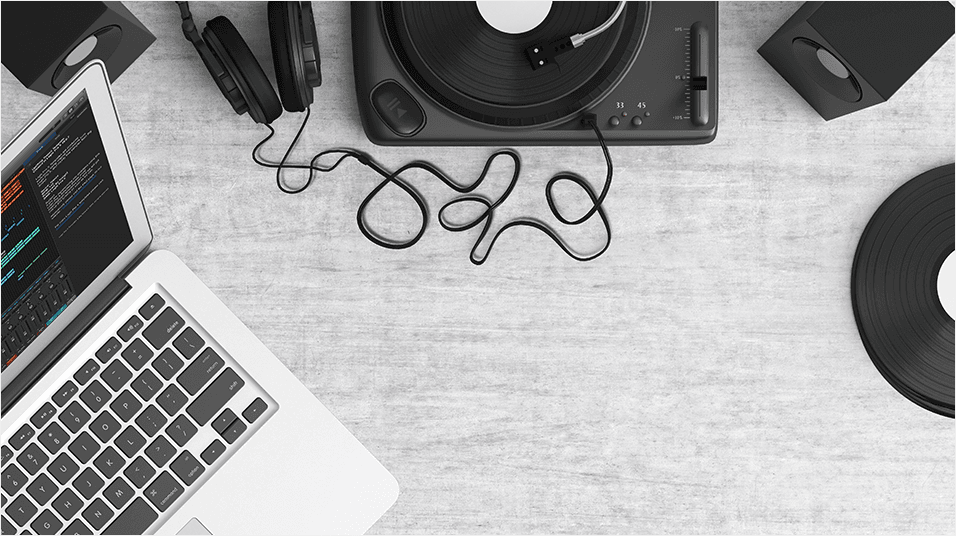 Against a grey backdrop, there is the corner of a silver Macbook on the left. On the right there is the side of a black record. In the centre there are some tangled wires.