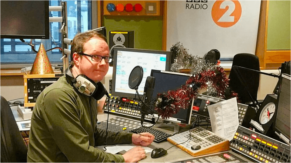 this is a picture of a white man with glasses, Tim Johns, looking at the camera. He sits in front of a laptop and a microphone at BBC Radio 2 studios.