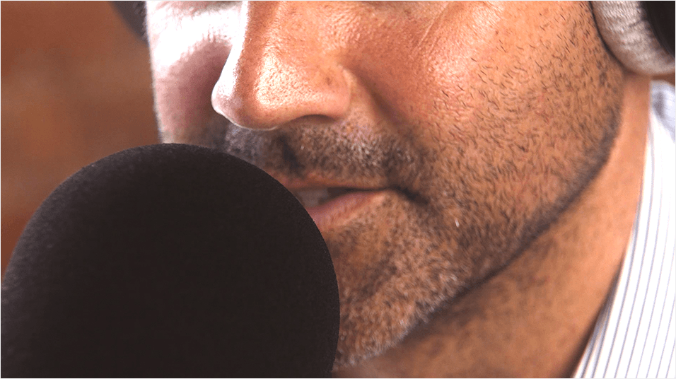 A close up of a white man with a closely shaven beard. He speaks into a rounded black microphone and has headphones on. We can't see anything above his nose.