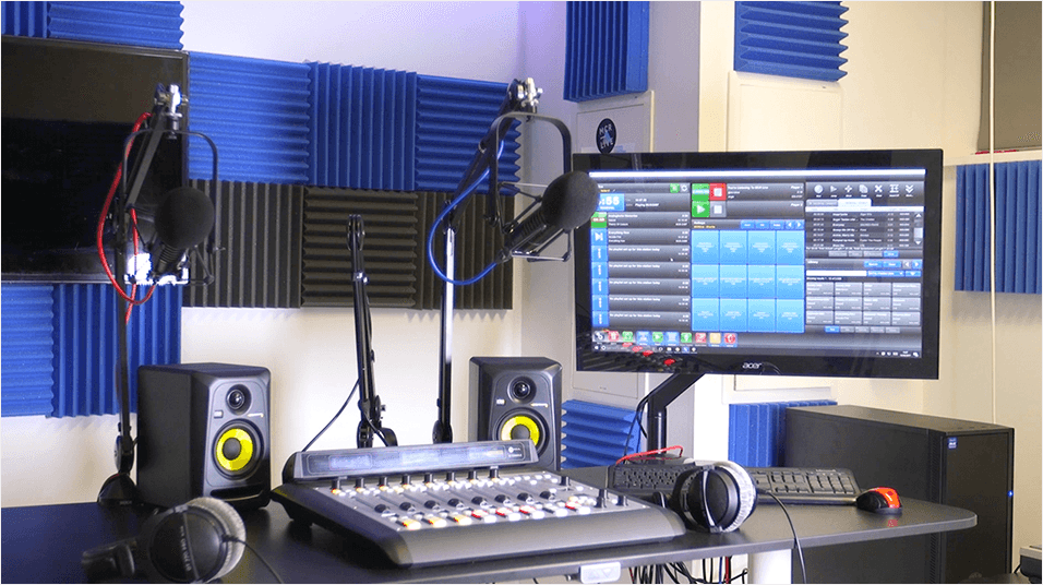 Image shows an online radio station studio: featuring a mixing console, two microphones, two sets of headphones, a computer and two speakers on a desk.